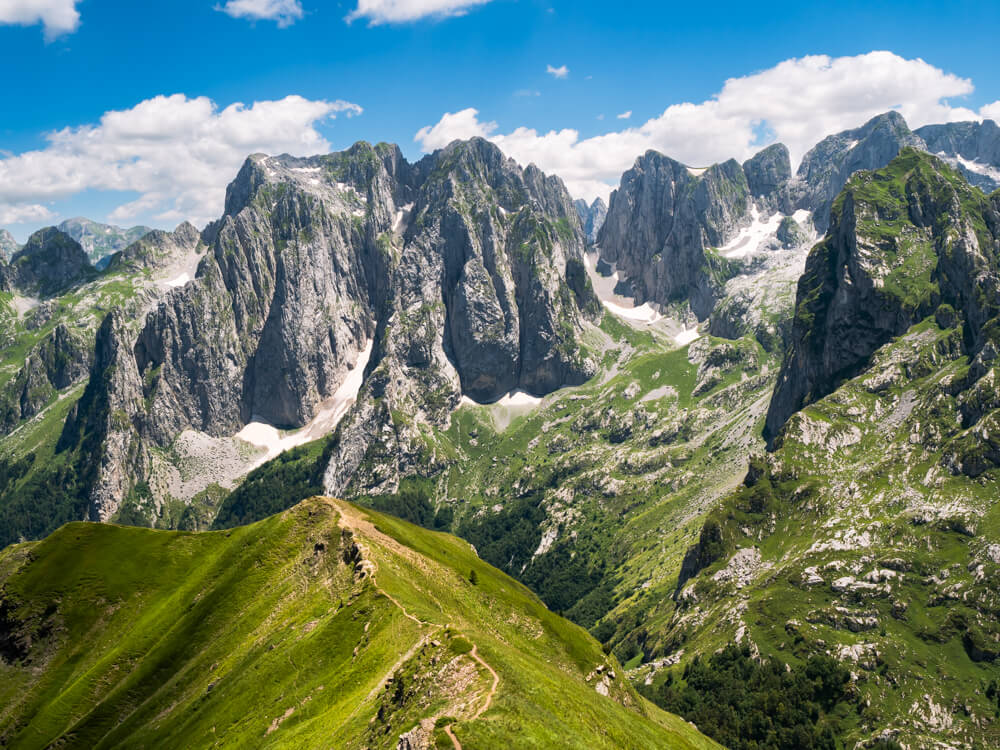 A panoramic view of rocky peaks with patches of snow and a trail along the ridge of a grassy mountain in the foreground, viewed from Maja e Vajushes, the best hike in the Albanian Alps.