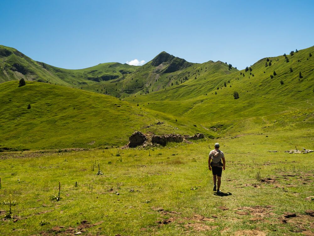 My husband walking across a green meadow at the bottom of a valley surrounded by mountains, with Vajusha Peak in the background.