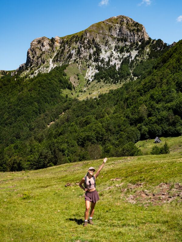 Me standing and waving on a green meadow, with a steep rocky peak towering behind me on our hike to Maja e Vajushes.