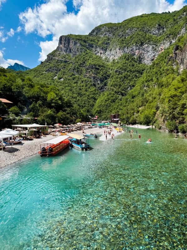 Turquoise Shala River surrounded by steep cliffs covered with green vegetation. The pebbly shores of the river are lined with sunbeds and umbrellas, and swimmers are wading in the crystal-clear water.
