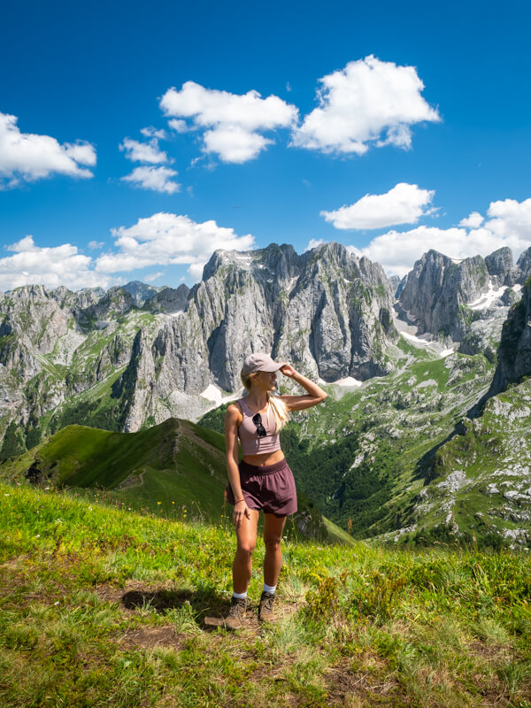 Me standing on the grassy Vajusha Peak, with vertical jagged limestone peaks in the background.